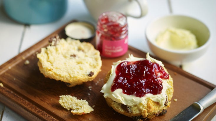 A scone is cut in half on a wooden board. One half is covered with cream, then jam! Behind it is the pot of jam and a light coloured cup containing tea, next to a blue ceramic teapot.